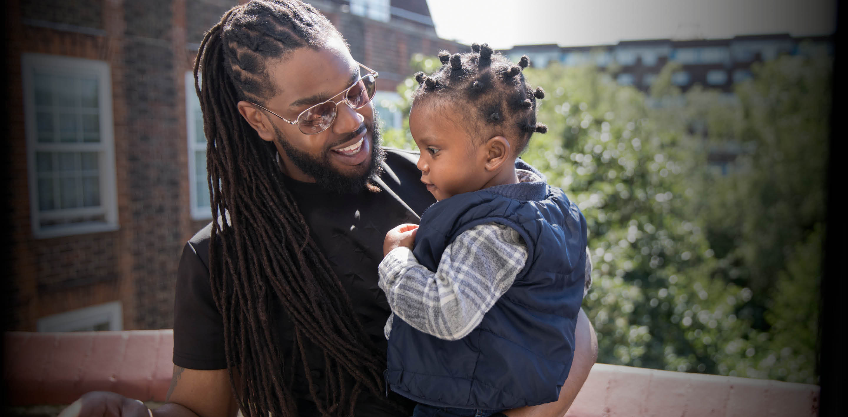Father with long braids holding toddler son on sunny balcony