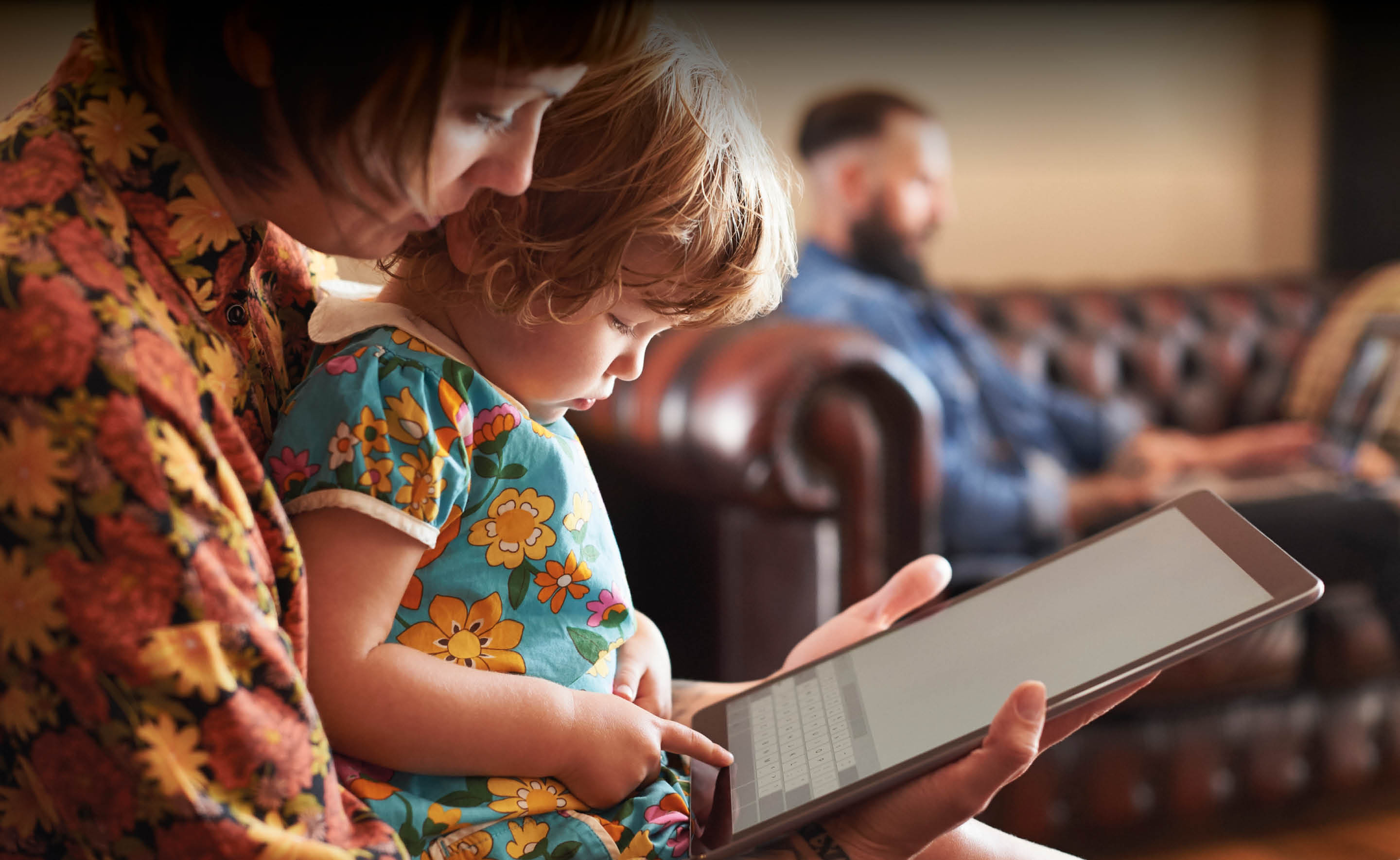 Transform : Customer Experience: A child sits on her mother lap and plays with a large electronic tablet. A father sits in the background on a laptop.